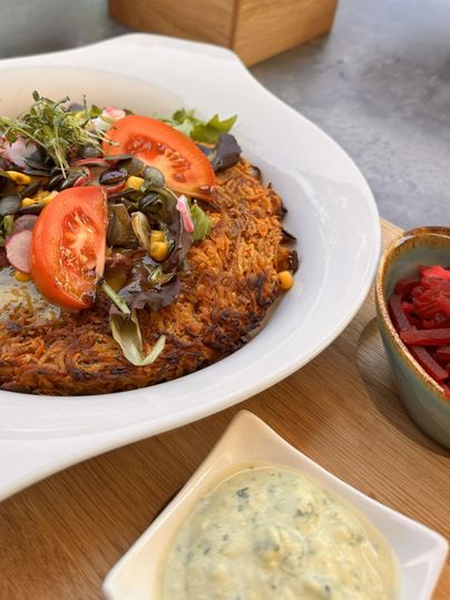 Ein herzhaftes Gericht aus geriebenen Kartoffeln, garniert mit frischem Gemüse und Tomatenscheiben. Daneben befinden sich zwei verschiedene Dips in Schalen.