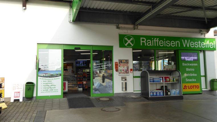 Ein Markt mit einem Eingangsbereich in grünen Farben. Es sind Werbetafeln und Regale sichtbar, die verschiedene Produkte anzeigen.