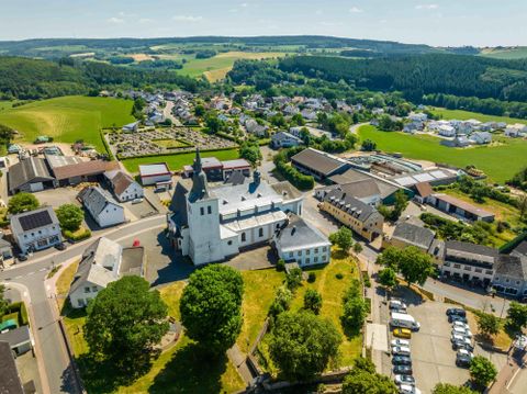 Eine idyllische Landschaft mit einer kleinen Kirche im Mittelpunkt. Im Hintergrund sind grüne Felder und Wohngebäude zu sehen.