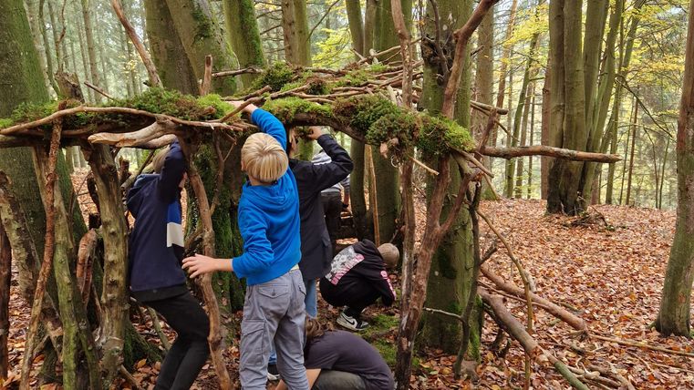 Eine Gruppe von Kindern baut eine Konstruktion aus Ästen und Zweigen im Wald. Die Umgebung ist herbstlich mit buntem Laub auf dem Boden.