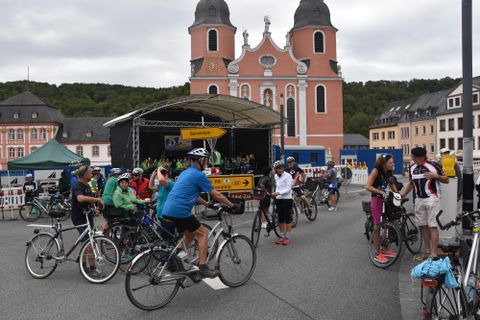 A group of cyclists gathers in front of a historic church. In the background, buildings and a stage can be seen.