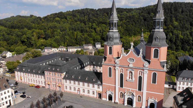 Eine beeindruckende Kirche mit zwei Türmen steht im Mittelpunkt des Bildes. Im Hintergrund sind grüne Hügel und ein gepflasterter Platz zu sehen.
