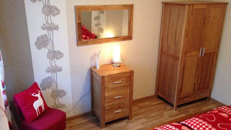 Ein gemütliches Schlafzimmer mit einem Holzkasten und einem Nachttisch. Die Wand hat ein florales Muster und es gibt ein rotes Kissen auf einem Sessel.