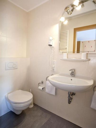 Ein modernes Badezimmer mit einer Toilette, einem Waschbecken und einem großen Spiegel. Die Wände sind neutral gefliest und der Boden hat eine graue Oberfläche.