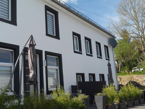 Ein modernes, zweigeschossiges Haus mit weißen Wänden und schwarzen Fensterrahmen. Vor dem Gebäude stehen Pflanzkübel mit grünen Pflanzen.