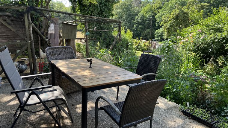 Ein gemütlicher Außenbereich mit einem Tisch und Stühlen, umgeben von Pflanzen und Bäumen. Der Himmel ist klar und sonnig.