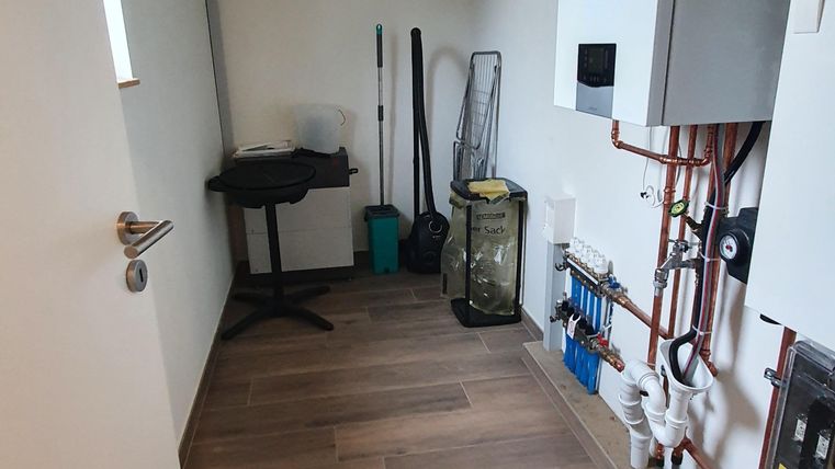 Ein schmaler Raum mit Holzdecke und warmen Fliesenboden. An der Wand sind Rohre und ein Heizgerät zu sehen, daneben steht ein Tisch.