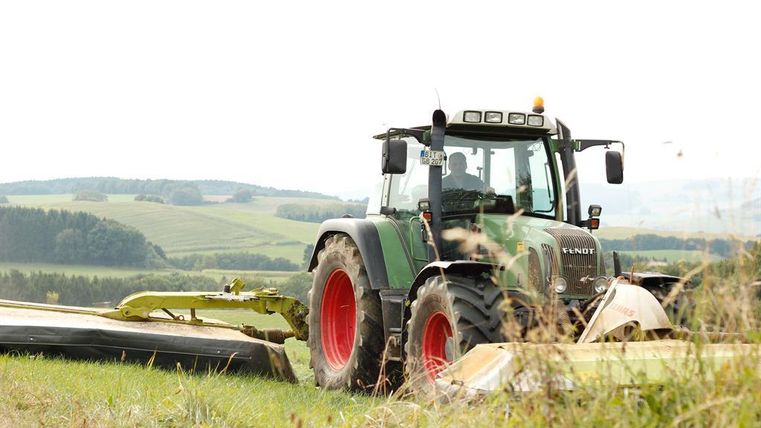 Ein grüner Traktor mäht ein Feld in einer ländlichen Umgebung. Im Hintergrund sind sanfte Hügel und Bäume zu sehen.