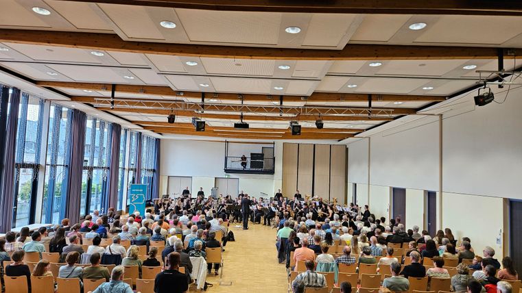 Une grande salle avec de nombreux sièges est remplie de public. Sur scène, un orchestre joue devant une vaste foule de spectateurs.