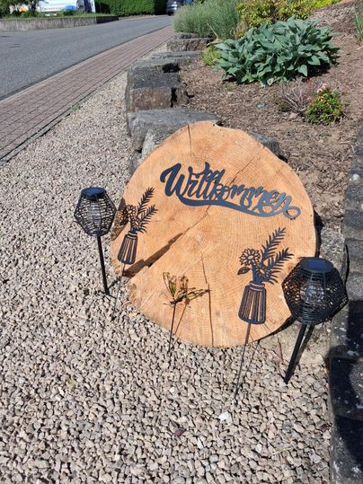 Ein dekoratives Holzschild mit der Aufschrift "Willkommen" steht auf Schotter. Neben dem Schild befinden sich zwei schwarze Laternen und etwas Grünpflanzen.