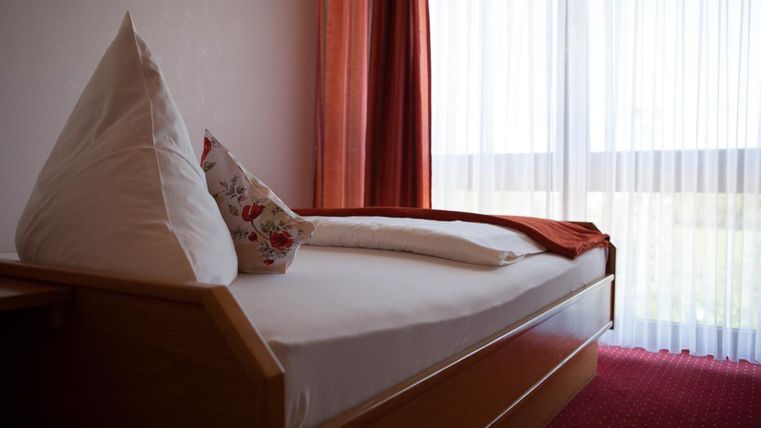 Ein gemütliches Zimmer mit einem Holzbett und flauschigen Kissen. Durch die großen Fenster strömt viel Tageslicht herein.