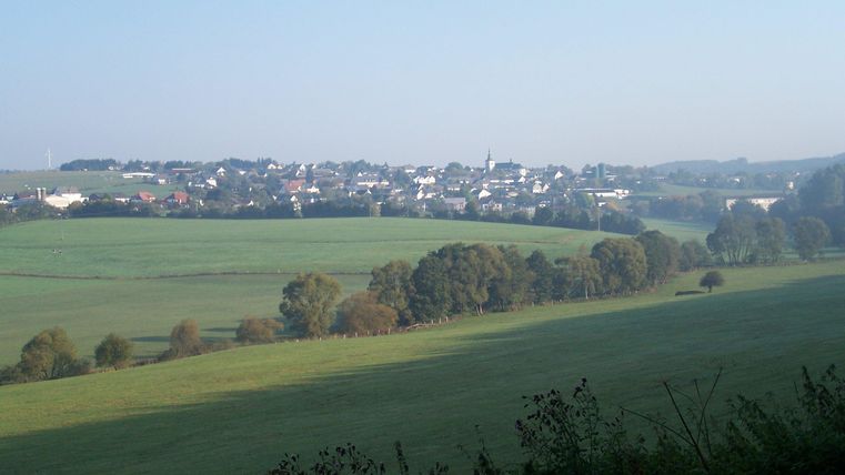 Eine ruhige Landschaft mit sanften Hügeln und weitläufigen Feldern. Im Hintergrund ist ein kleines Dorf mit weißen Häusern zu sehen.