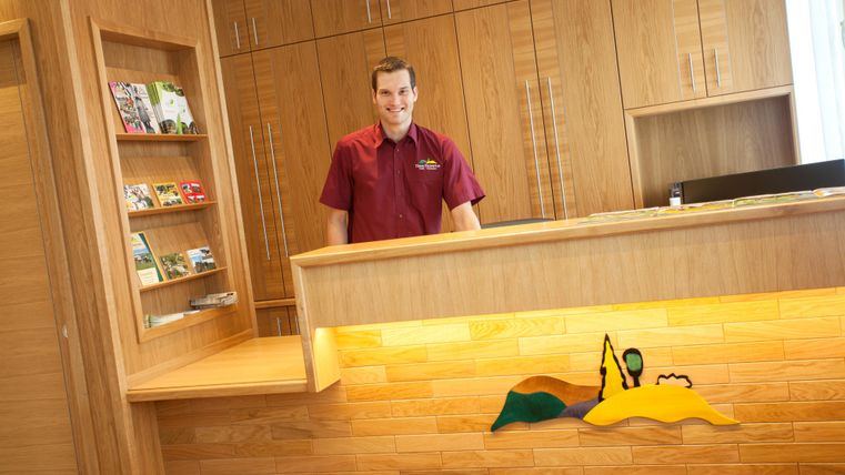 Ein Empfangsbereich mit einem freundlichen Rezeptionisten. Der Holzstil und die Regale mit Broschüren verleihen dem Raum eine warme Atmosphäre.