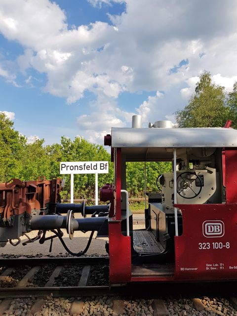Ein roter Zug steht auf den Gleisen an der Station Pronsfeld Bf. Der Himmel ist teils bewölkt mit einigen schönen Wolken.