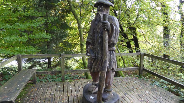 Eine Holzstatue eines Wanderers steht auf einer Plattform im Wald. Umgeben von grünen Bäumen und einer Naturkulisse.