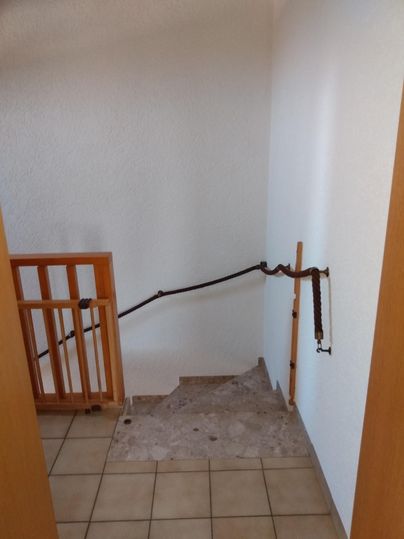 Ein Treppenabsatz mit einem Holzgeländer und einem geflochtenen Seil. Der Boden ist teilweise uneben und nicht ganz fertiggestellt.