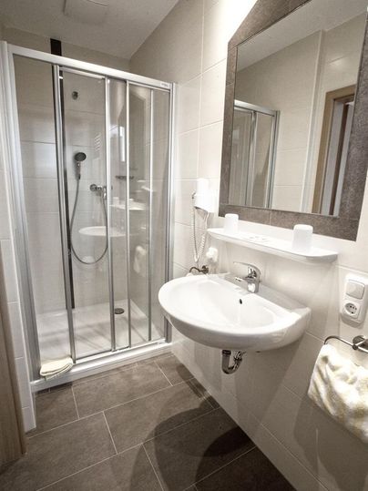 Ein modernes Badezimmer mit einer Dusche und einem Waschbecken. Die Wände sind hell gefliest und der Boden hat dunkle Fliesen.