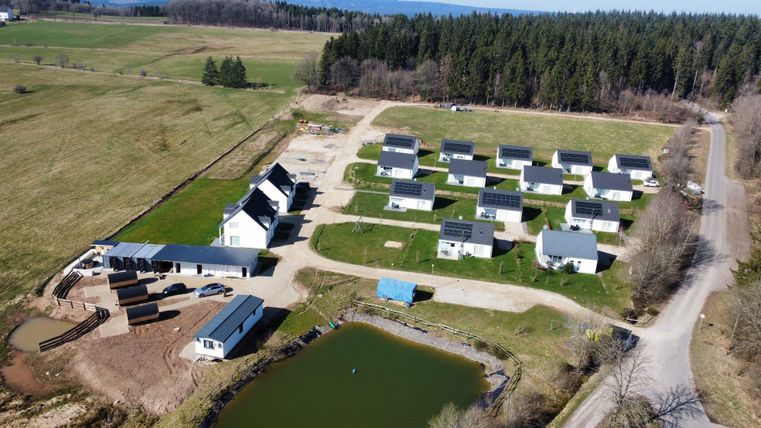 Eine Luftaufnahme einer Ferienanlage mit mehreren Hütten in einer grünen Umgebung. Im Vordergrund ist ein kleiner Teich und im Hintergrund erstrecken sich Wälder.