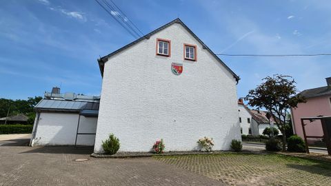 Ein einfaches, weißes Gebäude mit einem roten Wappen an der Wand. Im Hintergrund sind einige Bäume und andere Gebäude sichtbar.