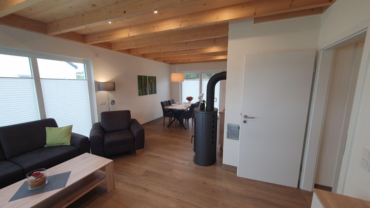 Ein modernes Wohnzimmer mit Holzdecke und großen Fenstern. Der Raum ist gemütlich mit einem Sofa, einem Tisch und einem Essbereich gestaltet.