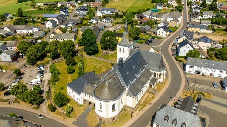 Een luchtfoto van een klein dorp met een kerk in het midden. De omgeving wordt omringd door groene velden en woningen.
