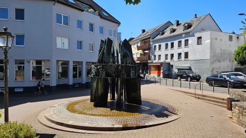 Ein modern gestaltetes Gebäude mit großen Fenstern und einem Glockenturm. Im Vordergrund ist ein begrüntes Gelände mit Bäumen und Straßenlaternen zu sehen.