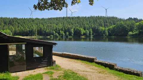 Ein ruhiger See umgeben von dichten Wäldern und Windkraftanlagen. Im Vordergrund ist ein Holzhaus sichtbar.