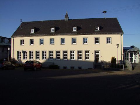 Ein mehrstöckiges, helles Gebäude mit einem schwarzen Dach und vielen Fenstern. Vor dem Gebäude stehen einige Autos und Beleuchtungssäulen.