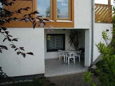 Ein gemütlicher Außenbereich unter einem Balkon mit einem Tisch und Stühlen. Grünpflanzen umgeben den Raum und sorgen für eine einladende Atmosphäre.