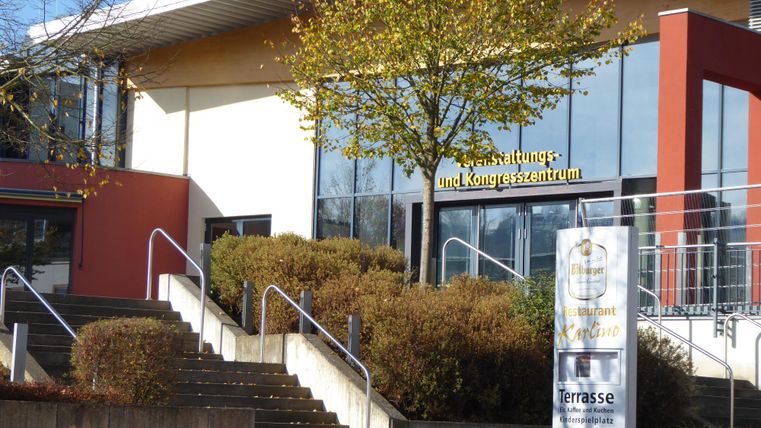 Ein modernes Veranstaltungs- und Kongresszentrum mit einer großen Fensterfront. Vor dem Gebäude befinden sich Treppen und eine gepflegte Grünanlage.