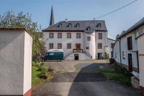 A historical building with a pointed tower and a large entrance front. In the foreground, there are a few vehicles and green landscapes.
