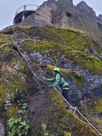 Eine Person klettert eine steile Felswand mit Seilunterstützung. Im Hintergrund sind Ruinen und eine Plattform sichtbar.