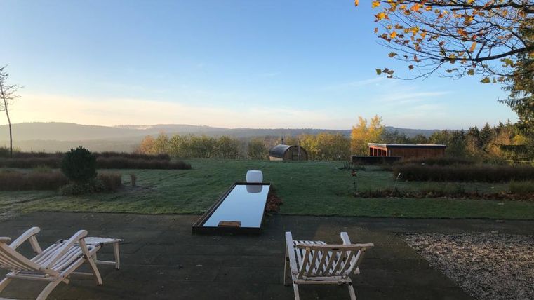 Eine ruhige Terrasse mit zwei Liegen und einem langen, spiegelnden Wasserbecken. Im Hintergrund erstreckt sich eine grüne Landschaft unter einem klaren Himmel.