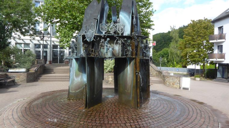 Eine moderne Brunneninstallation mit eleganten, metallischen Formen steht im Zentrum eines Platzes. Umgeben von Bäumen und Gebäuden, strahlt sie einen besonderen Charme aus.