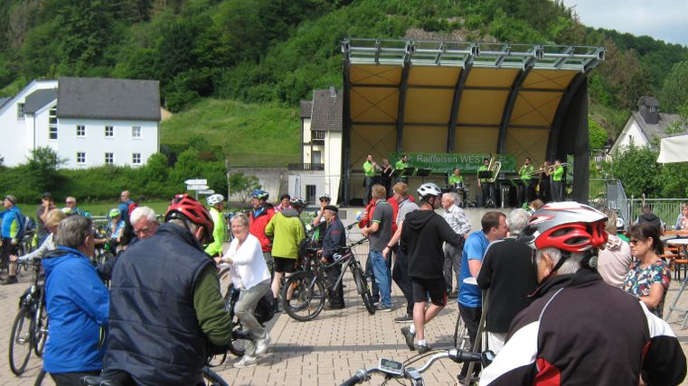 Eine Gruppe von Radfahrern versammelt sich auf einem Platz vor einer Bühne. Im Hintergrund sind grüne Hügel und Gebäude zu sehen.