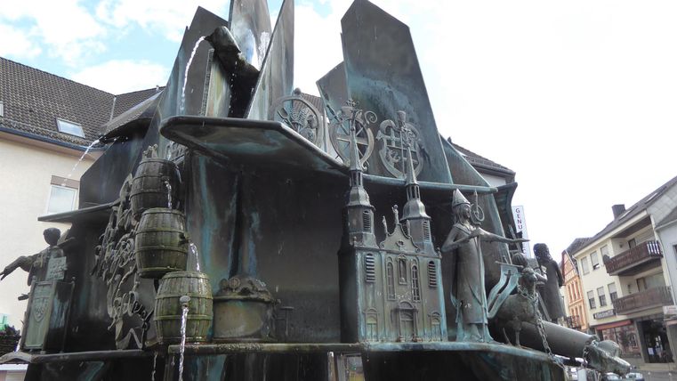 Ein kunstvoller Brunnen mit verschiedenen Skulpturen, der dynamische Figuren darstellt. Die Bronzeoberfläche reflektiert das Licht und schafft ein interessantes Spiel von Schatten und Form.