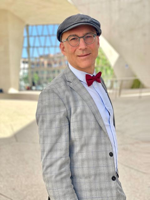 Ein stilvoll gekleideter Mann in einem grauen Anzug mit einer roten Fliege und einer Mütze. Er steht vor einer modernen Architektur im Hintergrund.