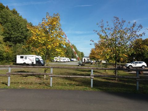Ein Campingplatz mit mehreren Wohnmobilen und Autos. Die Umgebung ist von Bäumen und grünem Gras umgeben, unter einem klaren blauen Himmel.