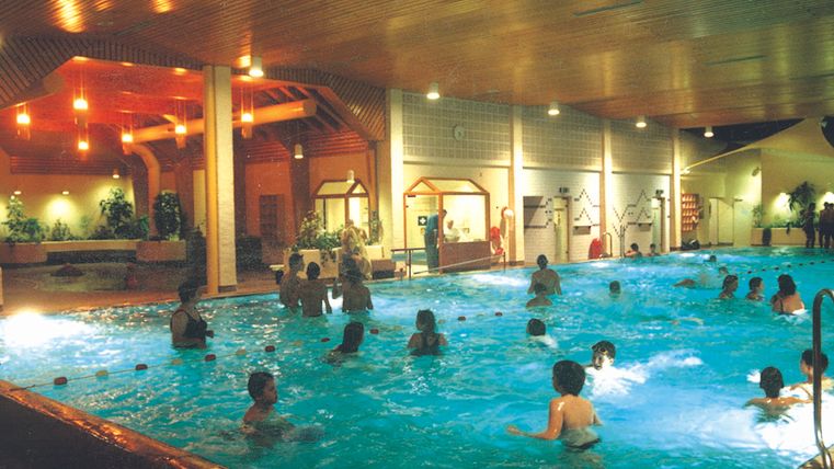 Ein Innenpool mit vielen Menschen, die im Wasser schwimmen und spielen. Die Umgebung ist hell erleuchtet und hat eine entspannte Atmosphäre.