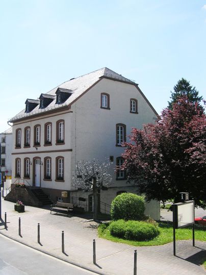 Ein gemütliches, zweistöckiges Haus mit weißen Wänden und einem grauen Dach. Vor dem Haus stehen grüne Bäume und hübsche Sträucher.