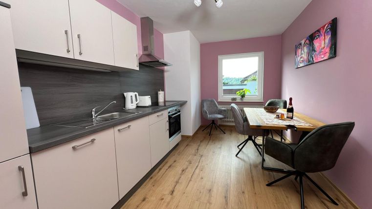 Eine moderne Küche mit hellen Möbeln und einer Essgelegenheit. Die Wände sind in sanften Farbtönen gehalten, und ein Fenster lässt viel Tageslicht herein.