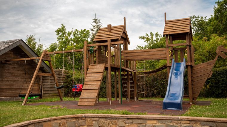 Ein Spielplatz aus Holz mit Rutschen, Leitern und Schaukeln. Die Umgebung ist grün und einladend.