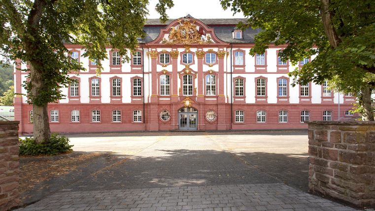Ein schönes historisches Gebäude mit roter Fassade und weißen Fenstern. Der Eingang ist von Bäumen umgeben und der Weg führt direkt zur Tür.