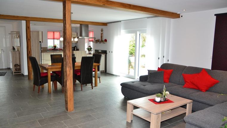 A modern living room with a comfortable gray couch and red cushions. The dining area with a wooden table and chairs is bright and inviting.