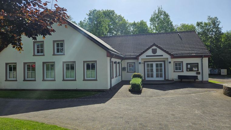 Ein modernes, zweigeschossiges Gebäude mit weißen Wänden und einem roten Dach. Es befindet sich in einer grünen Umgebung und wirkt einladend.