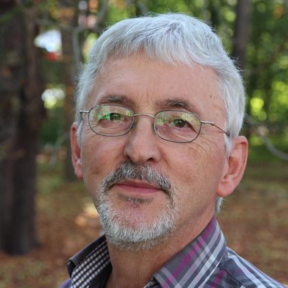 Ein älterer Mann mit grauen Haaren und Brille lächelt in die Kamera. Der Hintergrund ist unscharf und zeigt Bäume und herbstliche Farben.