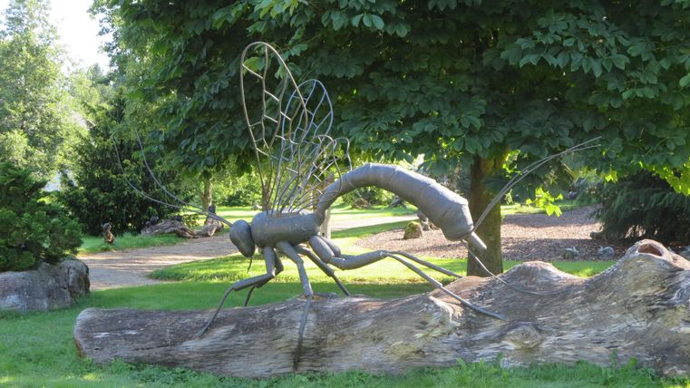 Eine große Metallskulptur eines Insekts sitzt auf einem Baumstamm. Die Umgebung ist grün mit Bäumen und einem gepflegten Garten.