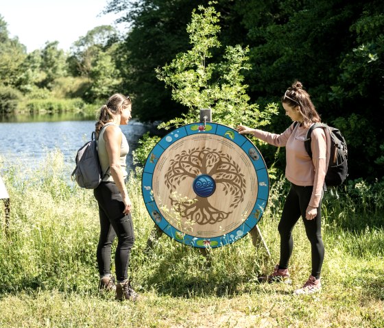 Zwei Frauen mit Rucksäcken betrachten ein dekoratives Holzrad am Ufer eines Sees, umgeben von grüner Natur., © Eifel Tourismus GmbH, Dominik Ketz