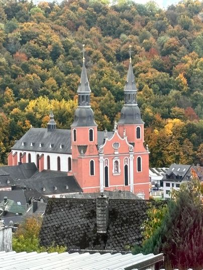 Eine Kirche mit zwei spitzen Türmen steht vor einem bunten Wald. Die Farben der Bäume zeigen den Herbst.