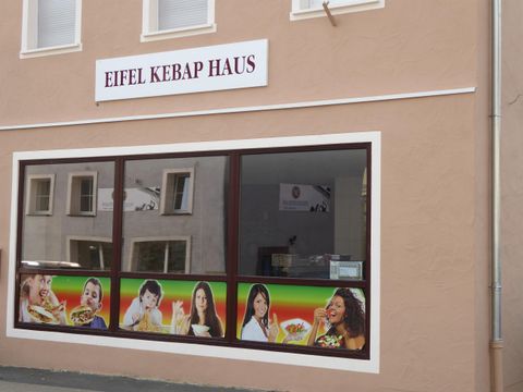 Un restaurant nommé "Eifel Kebap Haus" avec de grandes fenêtres. La façade montre des images de différents plats et de personnes joyeuses.